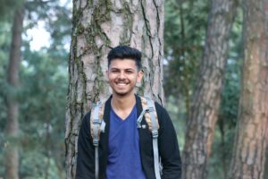 Young man in blue shirt with a backpack on in front of a tree.