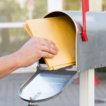 A hand putting a manila envelope into a mailbox with a red flag.