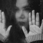 A sad looking woman in black and white presses her hands against a window pane as it rains