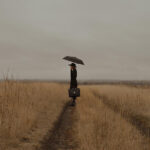 A photo of a woman holding an umbrella stands alone in a field.