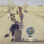 A young woman sits on a crate looking at a map. Next to her is a suitcase and a globe.